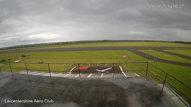 A view from a webcam at Leicestershire Aero Club shows the tarmac runway and a helicopter landing pad with a white rotor symbol. The sky is overcast with dark, heavy clouds, and the surrounding grassy fields stretch out into the distance. A small flag is visible on the left side near a pole, and there are some signs and markings on the grass. The timestamp indicates the date as July 6, 2025, at 14:00.