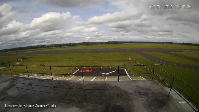 A view from a webcam at Leicestershire Aero Club showing a grassy airfield with taxiways and runways stretching into the distance under a partly cloudy sky. In the foreground, there is a helicopter landing pad with a large white "X" marked on it, surrounded by a black fence. An emergency wind sock is visible on the left side. The timestamp indicates the date and time as July 6, 2025, at 15:00:25.