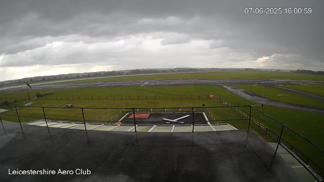 A view from a webcam at Leicestershire Aero Club shows an outdoor scene under a cloudy sky. In the foreground, there is a helipad with a large helicopter rotor blade resting on it. Behind the helipad, a grassy area is enclosed by a small fence, and a windsock is visible on a pole. The surrounding landscape consists of open fields, some covered with what appears to be water or reflective surfaces, and a few structures in the distance. The timestamp in the upper right corner indicates the date and time as July 6, 2025, at 4:00 pm.