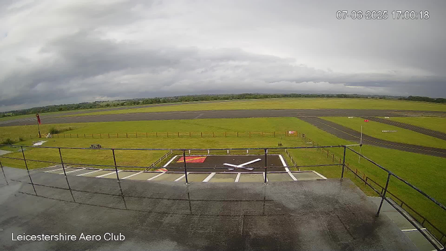 An outdoor view from a webcam at Leicesterhire Aero Club showing a cloudy sky over a grassy airfield with runways and taxiways. In the foreground, there is a helipad marked with a white "H" on a red background, enclosed by a black railing. The landscape is flat with a mix of green grass, paved runways, and distant trees. The scene appears to be during daytime with overcast weather. The timestamp on the image indicates the date and time as July 6, 2025, at 17:00:18.