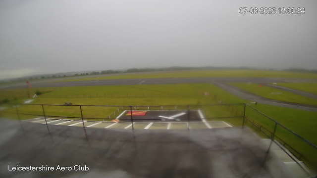 A view from a webcam at Leicesterhire Aero Club shows a cloudy sky over an empty runway and grassy areas. A small section of the observation deck, with railings and parking spaces, is visible at the bottom of the image. The date and time in the top right corner read "07-06-2025 18:00:24."