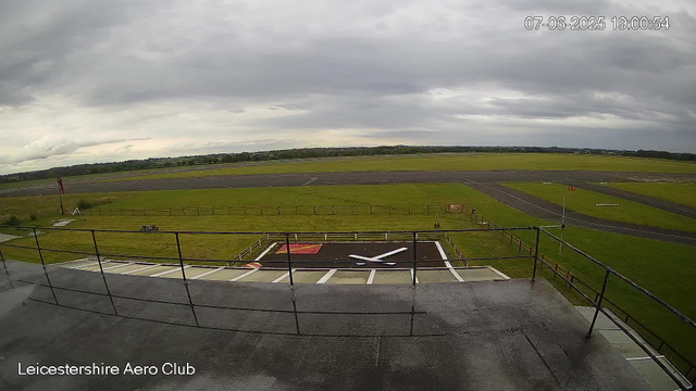 A view from a webcam at Leicestershire Aero Club shows an overcast sky with dark clouds. The image includes a small helipad with a large white "H" symbol, surrounded by a railing, in the foreground. The tarmac area behind the helipad extends into grassy fields with taxiways and runways visible in the distance, all under gray, cloudy skies. The timestamp in the upper right corner indicates the date as July 6, 2025, and the time as 19:00:54.