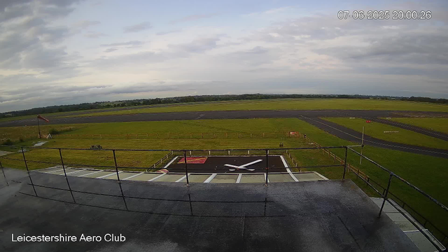 An outdoor view from a webcam at Leicestershire Aero Club showing a cloudy sky, a grassy airfield with a small landing area marked by runway lines, and a windsock on the left side. The image is taken from a rooftop or elevated platform, with a railing in the foreground. The timestamp indicates the date and time as July 6, 2025, at 8:00 PM.