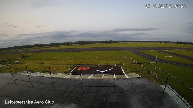 A view from a webcam at Leicestershire Aero Club showing a runway and surrounding grassy area under a cloudy sky. There is a helicopter landing pad with a large clock painted on it in the foreground. The timestamp on the image indicates it was taken on July 6, 2025, at 21:00:23. An orange windsock is visible on the left side of the image. A fence surrounds the helicopter pad, and traffic cones mark the edges of the runway. The landscape extends to a horizon of fields and distant trees.