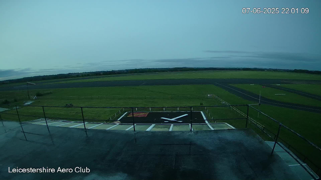 An empty helicopter landing pad with a large white "X" symbol in the center, surrounded by a black border. The landing pad is on a flat, open grassy area with a few small structures and equipment nearby. In the distance, there are open fields and a cloudy sky. The bottom left corner has the text "Leicestershire Aero Club." The date and time in the top right corner shows "07-06-2025 22:01:09."