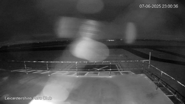 A black-and-white image from a webcam shows a dark night scene with a parking area and a safety railing in the foreground. There are blurred light spots in the air, possibly raindrops or moisture on the camera lens. The background features an open, flat landscape with a few distant lights on the horizon. The timestamp in the upper right corner reads "07-06-2025 23:00:36," and the bottom left corner displays "Leicestershire Aero Club."