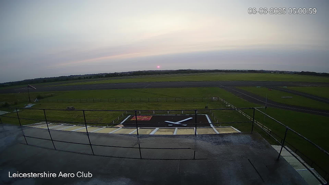 An early morning view from a webcam at Leicester Aero Club. The horizon shows the sun just beginning to rise, casting a faint pink glow. The scene features a grassy airfield with a large clock painted on the ground below the camera, displaying the time as approximately 5:00. The date and time are displayed in the top right corner as June 8, 2025, at 5:00:59. The airfield's runways extend into the distance, surrounded by open fields under a partly cloudy sky.