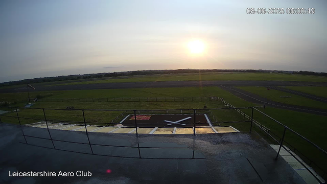 The image shows a view from a webcam at Leicestershire Aero Club during sunset or sunrise. In the foreground, there is a large black and white ground marking of an airplane silhouette on a paved area, surrounded by metal safety railings. Beyond this, there is a grassy area with a small flagpole and a windsock. In the background, the airfield extends with runways and taxiways visible, set against a horizon of distant trees and open sky. The photo is timestamped "08-06-2025 06:00:49" in the top right corner.
