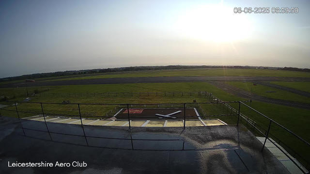 An outdoor view of a runway at Leicester Aero Club during sunrise. The sky is mostly clear with the sun shining brightly near the horizon. In the foreground, there is a black helipad with a white helicopter rotor symbol, surrounded by a fence and some tables or equipment. The grassy area surrounds the runway and extends into the distance, with a few signs and markings visible near the runway. The timestamp indicates the photo was taken on June 8, 2025, at 6:59 AM.