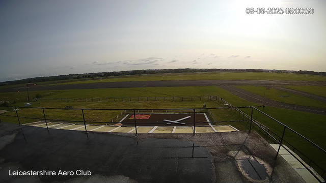A view from a webcam at Leicestershire Aero Club showing an airfield with a runway, a grassy area, and a large clock painted on the ground in the foreground. The sky is partly cloudy with the sun shining, and there is a windsock visible on the left side. The timestamp in the top right corner reads "08-06-2025 08:00:30".