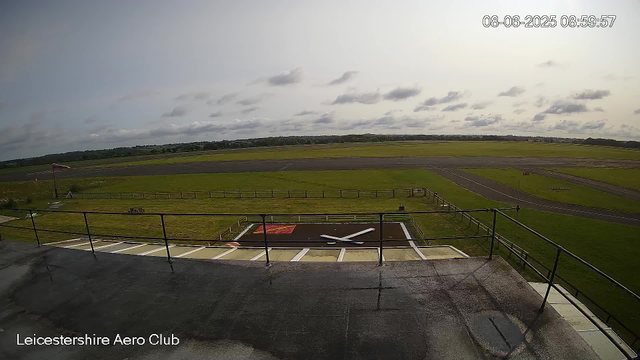 An empty tarmac runway with a small helipad marked by a white helicopter silhouette, situated in front of a grassy field. A wind sock is visible on the left side, and a wooden fence encloses part of the area. In the background, there are open grassy fields and a cloudy sky. The timestamp in the top right corner reads "08-06-2025 08:59:57," and the bottom left corner has the text "Leicestershire Aero Club."