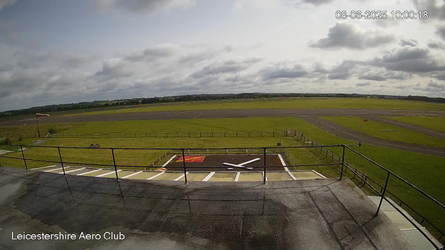 A wide view of Leicesterhire Aero Club's airfield under a cloudy sky, showing a small helipad with a white helicopter silhouette painted on it in the foreground. The control tower or observation deck railing is visible at the bottom of the image, with the club's name in the bottom left corner.