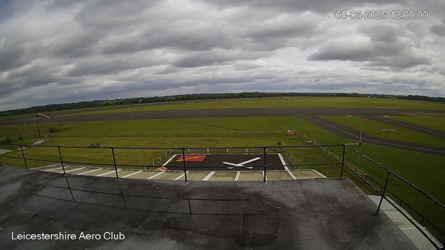A wide view from a webcam at Leicesterhire Aero Club shows a cloudy sky over a grassy and paved area. In the foreground, there is a large helicopter landing pad with a white helicopter silhouette painted on it. The scene includes a metal railing and a flagpole with a flag on the left side. The background features an open field, a few small structures, and a runway or taxiway running horizontally across the image. The timestamp on the image reads "08-06-2025 12:00:00".