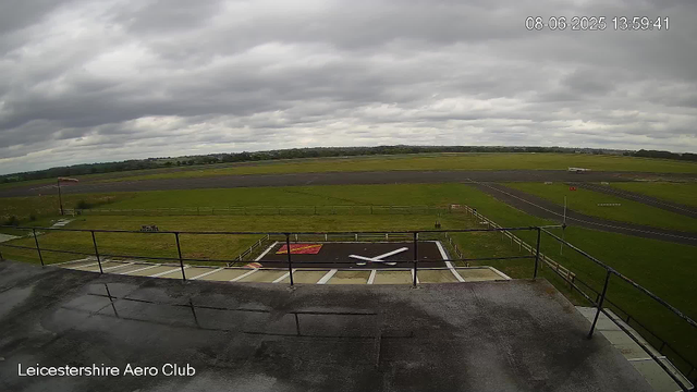 An empty rooftop observation area at Leicestershire Aero Club overlooks an airport runway under a cloudy sky. The clock on the helipad reads 13:59.41 and the date is August 6, 2025. An airplane is visible on the runway in the distance.
