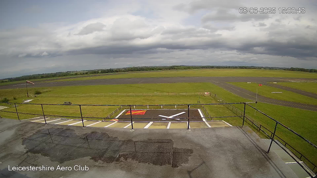 The image shows an outdoor view of a small aircraft helicopter landing pad at Leicestershire Aero Club. The landing pad has a large "H" marking in white in the center, surrounded by white lines and a red square. A weather vane with a flag is situated near the pad on the left side, and there are fences enclosing the area. Beyond the fenced area, there are green fields, taxiways, and open land extending towards the horizon. The sky is mostly cloudy with some patches of blue. The timestamp indicates the date is June 8, 2025, at 15:00:43.