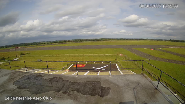 A view from a webcam at Leicestershire Aero Club showing an airfield with a large open grassy area, runways, and a small helipad. The sky is partly cloudy with patches of blue, and a flag is flying on a flagpole near the edge of the grassy area. There is a picnic table and some small structures or signs near the grassy area. The date and time displayed on the image are 08-06-2025 at 15:59:59.