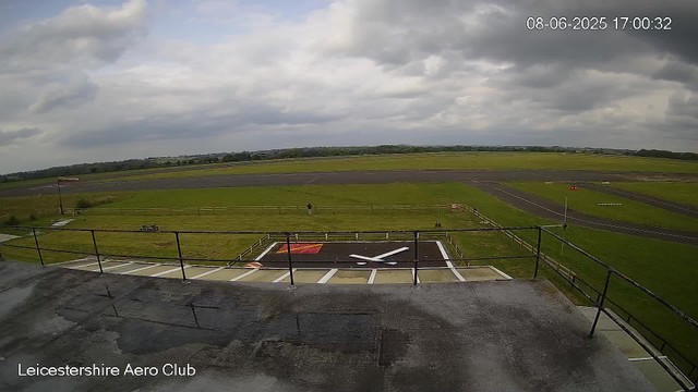 An overcast sky with dark clouds looms over Leicester Aero Club's runway and grassy airfield. In the foreground, a helipad marked with a large white "H" and a red square is visible, surrounded by a black railing. A small flag on a pole is positioned near the helipad, and a person stands near the fence on the grassy area. The runway extends into the distance, with multiple taxiways and navigational markers. The landscape is flat with some distant trees and low hills on the horizon. The timestamp reads "08-06-2025 17:00:32."