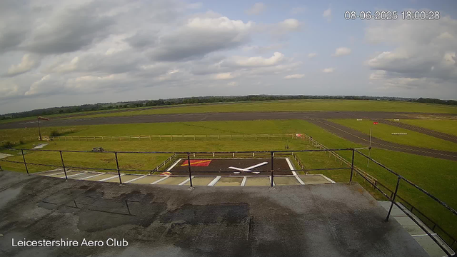 A view from a webcam at Leicesterhire Aero Club shows an empty helicopter landing pad with a large white "H" painted on a red background. The landing pad has black asphalt with white lines marking its boundaries. Surrounding the pad is a grassy area with a small wooden picnic table and bench. In the background, there is a wide open grassy field extending to the horizon, with a runway visible further back. The sky is partly cloudy with large, fluffy clouds scattered across a blue sky. A timestamp indicates the date as August 6, 2025, and the time as 18:00:28.