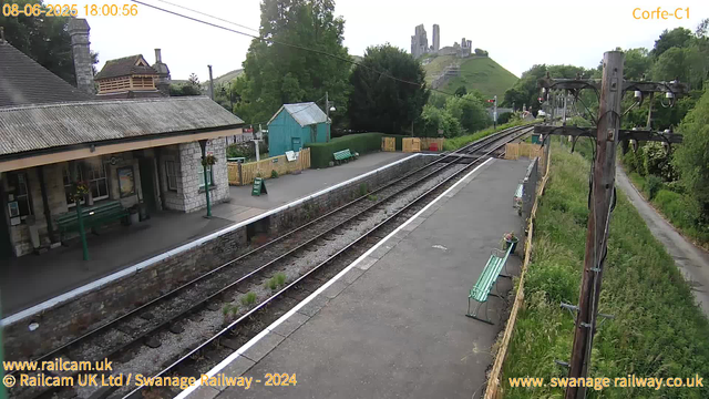 A small train station platform with a building on the left, featuring a tiled roof and stone walls. There are two green benches and a small turquoise shed nearby. The railway tracks run through the center, with a fence running alongside the platform. In the background, a hilltop with the ruins of a castle or fortress is visible, surrounded by lush green trees. Wooden utility poles with wires extend along the right side of the scene, and there is a narrow path with grass and bushes on the far right. The date and time are displayed at the top left corner.