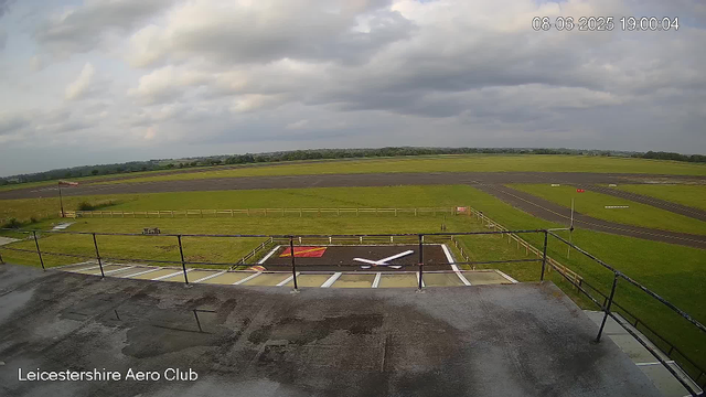 An aerial view of an airfield shows a grassy area, taxiways, and runways under a cloudy sky. The image includes a clock tower with a clock face, railway track, a small flag, and a fenced area. The timestamp indicates the date and time as June 8, 2025, at 7:00 PM. The location is identified as Leicestershire Aero Club.
