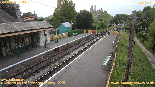 A small train station platform with a stone building on the left, green benches, and a teal shed in the background. Train tracks run through the center, with a rustic wooden fence separating the platform from green foliage. In the distance, a hill with ruins of a castle is visible under a partly cloudy sky. An electrical pole with wires stands on the right side. The image is timestamped "08-06-2025 20:00:38."