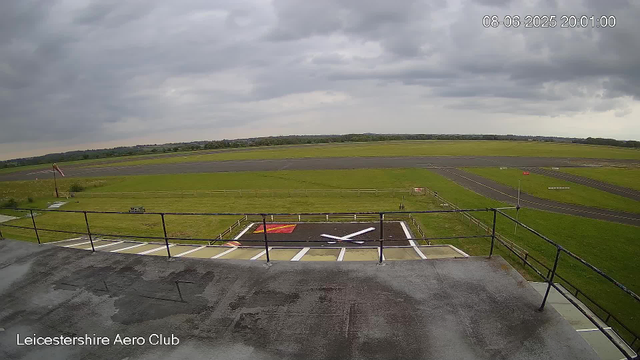 A wide view of an airport runway and taxiway under a cloudy sky. The foreground shows a rooftop or platform edge with a safety railing, and a helipad with a white helicopter symbol painted on it. A small windsock is visible on the left side, blowing towards the right. The landscape is predominantly grassy with some distant trees and low hills in the background. The timestamp in the top right corner reads "08-06-2025 20:01:00," and the bottom left corner indicates "Leicestershire Aero Club."