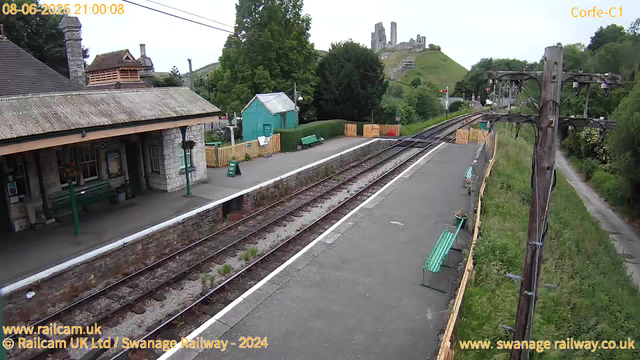 A small train station platform with empty benches, a building with a stone wall, and a green shed in the background. Railroad tracks run through the scene, with a hill and castle ruins visible in the distance. There are some wooden fences and electrical poles along the track. The time stamp shows "08-06-2025 21:00:08." Overcast sky and greenery surround the area.