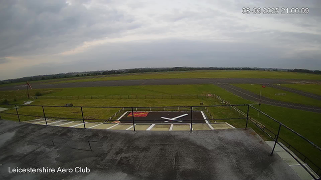 An overcast sky looms over an empty airport runway and taxiway at Leicestershire Aero Club. In the foreground, there is a red and white helicopter landing pad with a white "X" mark on it, surrounded by a black railing. The taxiway and runway stretch out into the distance with green grass on either side. The horizon shows a line of trees and distant fields under a cloudy sky. The timestamp indicates the date and time as August 6, 2025, at 21:00.