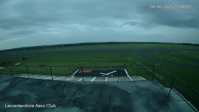 A view of an airfield taken from a webcam, showing a large helipad in the foreground with a white helicopter symbol painted on it. There is a balance beam in the distance, along with green grassy fields and a runway extending into the horizon under a cloudy sky. The image is labeled "Leicestershire Aero Club" in the bottom left corner and the timestamp reads "08-06-2025 22:00:02" in the top right corner.