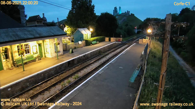 A small train station platform is shown in the evening with artificial lighting. There are two benches, a green bench on the left under a windowed building with hanging flower baskets, and another green bench along the platform next to a small turquoise shed. An "Away Out" sign is visible near the shed. To the right, there is an old wooden electric pole with mounted lights, supported by multiple wires. The railway tracks run through the center of the image and extend into the distance, curving slightly to the right. On the far side of the tracks, there is a grassy hill with trees and ruins on top, illuminated against the darkening sky. In the background, on the hill, there are remnants of structures that resemble castle ruins. The timestamp in the top left corner reads "08-06-2025 22:00:20".