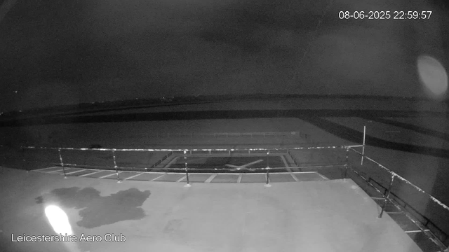 A black-and-white image captured by a webcam at night, showing an empty parking lot with several marked parking spaces and a few benches along the perimeter. There's a railing around a small, circular area in the center. The sky is dark, and the scene is illuminated by a faint light, possibly from the moon or distant streetlights. The text in the bottom left corner reads "Leicestershire Aero Club," and the top right corner displays the date and time: 08-06-2025 22:59:57.
