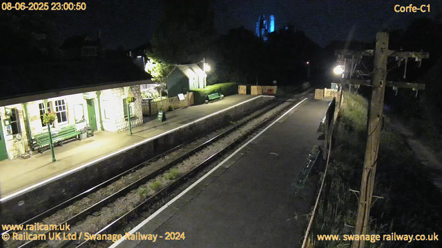 A quiet railway station platform at night, illuminated by a few lamp posts. To the left, there is a small building with white stone walls, green window frames, and flower baskets hanging outside. A green bench and a small shed are also visible. The railway tracks run through the center, with some grass growing between the rails. There is a wooden fence and some equipment on the right side, along with a utility pole carrying power lines. The scene is dark, with most of the area barely visible beyond the platform's illumination. The timestamp indicates the date is August 6, 2025, at 11:00 PM.