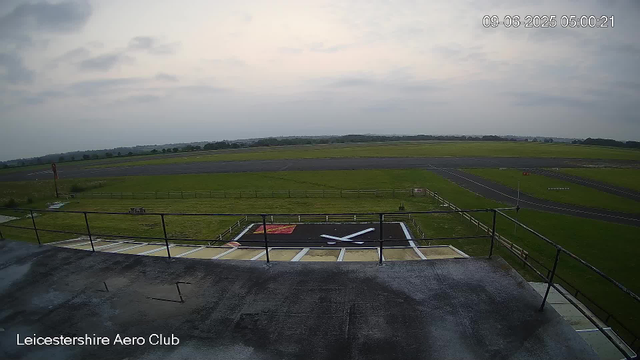 An outdoor view from a webcam at Leicestershire Aero Club. In the foreground, there is a helicopter landing pad with a black surface featuring a white helicopter symbol and a red border. The helipad is surrounded by a metal railing. Beyond the helipad, there are paved taxiways and grassy areas. In the distance, there are open fields and a few scattered trees under a cloudy sky. The timestamp on the image shows it was taken on September 6, 2025, at 5:00 AM.