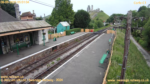 A small train station platform with a stone building on the left, decorated with hanging flower baskets. There are green benches along the platform, one near a "Way Out" sign. The train tracks run through the middle, with some weeds growing between the rails. There is a green wooden shed and a fenced area behind the platform, surrounded by trees and bushes. In the background, a hill with an old castle ruin sits atop a grassy slope. An old wooden telegraph pole with wires extends along the right side of the image, with greenery growing alongside a narrow pathway. The timestamp shows the date as June 9th, 2025, at 5:01 AM.