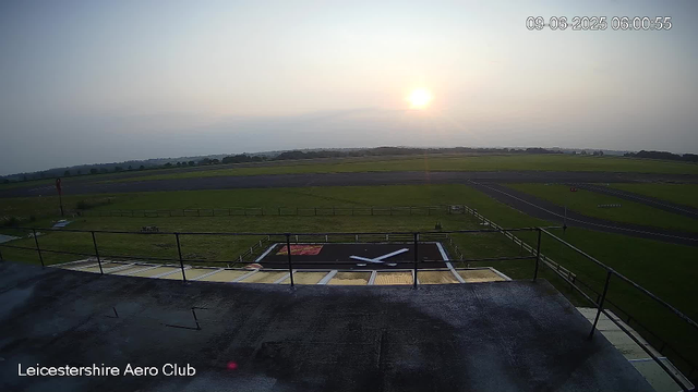 A view from a webcam at Leicesterhire Aero Club showing an early morning scene with the sun rising in the sky. The image includes a large runway in the distance, a grassy area in the middle, and a small helicopter landing pad with a white and black design in the foreground. The timestamp on the image indicates it was taken on September 6, 2025, at 6:00 AM. Some equipment and barriers are visible around the landing pad. The logo of Leicesterhire Aero Club is displayed at the bottom left corner.