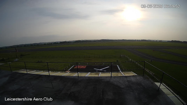 An aerial view of a runway and green fields at Leicestershire Aero Club during sunrise or sunset. The sky is partially cloudy with the sun low on the horizon. In the foreground, there is a helipad with a large white "X" on a black background, and a flag on a pole to the left. The image is timestamped "09-06-2025 06:59:52."