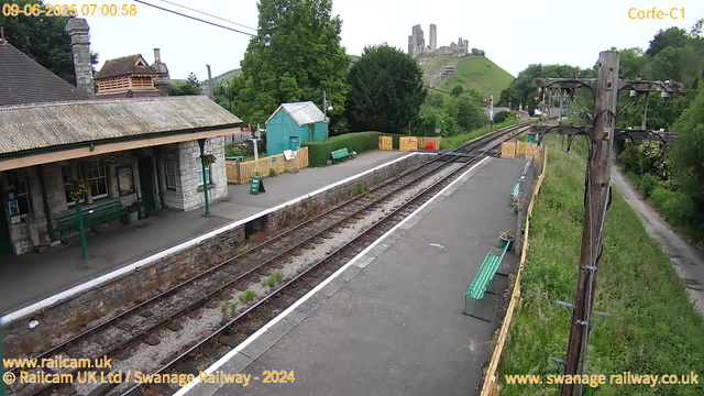 A small train station platform with two green benches, a small green shed, and a yellow fence. Overhead, there are electrical power lines. To the right, there's a gravel path running alongside the station. In the background, a hill with ruins or an old castle is visible, surrounded by lush green trees. The station building is on the left with stone walls and a tiled roof. The image is timestamped "09-06-2025 07:00:58" in the top left corner.