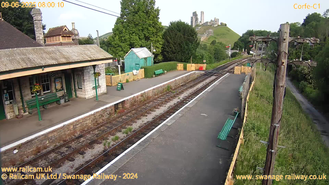 A small train station platform with a stone building, green benches, and flower baskets. There are railway tracks running alongside the platform, with a tall wooden pole holding electrical wires. In the background, a green hillside with a historic castle or ruins on top. The time and date are displayed at the top left corner as "09-06-2025 08:00:10," and the station name "Corfe-C1" is at the top right corner. The scene is outdoors on a clear day with lush green trees surrounding the area.