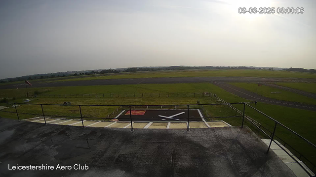 An empty helicopter landing zone with a painted red and black helicopter symbol on the tarmac, surrounded by grassy fields and a small fence, underneath a bright, partly cloudy sky. The image is labeled "Leicestershire Aero Club" at the bottom left corner and has a timestamp in the top right corner showing "09-06-2025 08:00:08".
