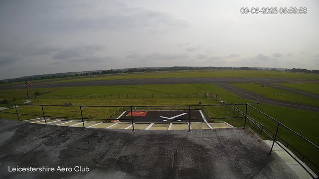 A view from a webcam at Leicestershire Aero Club showing an overcast sky, a grassy airfield with taxiways, and a small helipad with a white helicopter silhouette painted on it. The timestamp in the top right corner reads "09-06-2025 08:59:58."