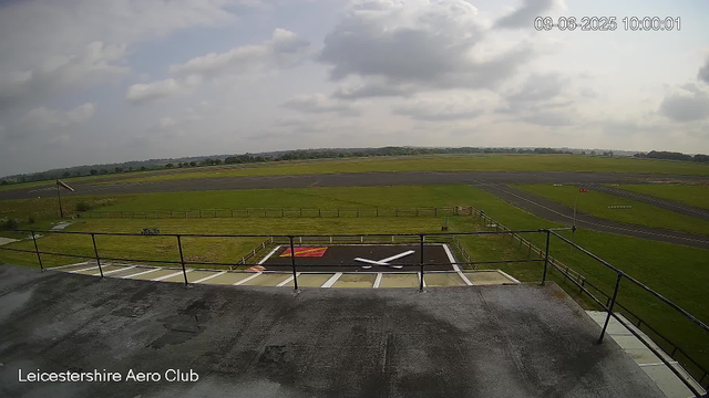 A view from a webcam at Leicestershire Aero Club showing an open green landscape with an airstrip in the distance, a helipad marked with a white "X" and a red square, surrounded by a safety fence. The sky is partly cloudy with patches of sunlight. The image includes a timestamp in the top right corner indicating it was taken on September 6, 2025, at 10:00:01.