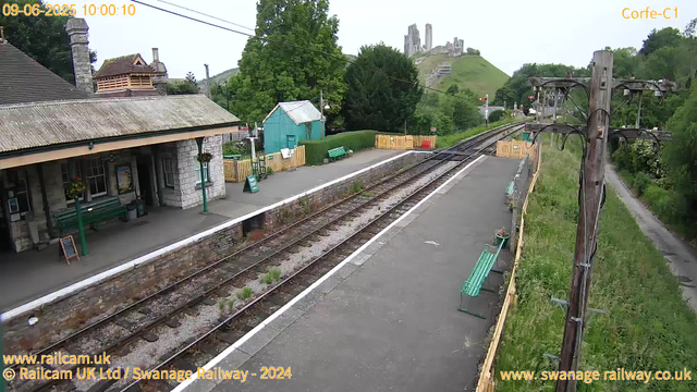 A small railway station platform with benches and a shelter, adjacent to railway tracks. In the background, there are trees, a green shed, a yellow fence, and a castle on a hill. A wooden utility pole with wires runs along the right side of the platform. The sky is overcast. The image has timestamps and website URLs superimposed on it.