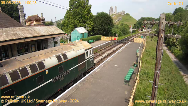 A vintage train car, painted green and cream, is stationary at a small railway platform labeled "Lly Jenny Lewis RN." The station has a stone building with a tiled roof on the left side and a small green wooden shed further back. The platform is bordered by a yellow safety barrier, and there are several green benches along its length. Overhead, electrical wires run parallel to the train tracks. Behind the station, lush green trees and a grassy hill are visible, with an ancient stone castle ruin perched atop the hill in the background. On the right side, there is a wooden utility pole with wires, and a narrow grassy path runs alongside the railway. The overall scene is bright and sunny, with clear skies and gentle green surroundings indicating a peaceful rural location. The timestamp indicates the image was captured on June 9, 2025, at 11:00:28 am.