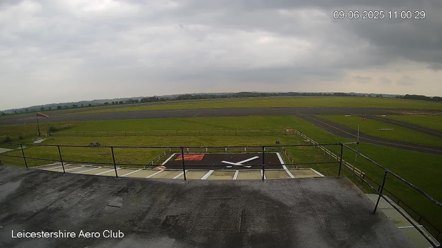 An open air section of Leicesterhire Aero Club shows a large grassy field surrounded by an asphalt runway and taxiways. In the foreground, a helipad with a white and red marking and an ‘X’ pattern is visible, along with a safety railing and a dark, slightly wet surface. The sky is overcast with gray clouds, and the landscape in the background features rolling hills and scattered trees. There is a small red and white windsock visible on the left side of the image. The timestamp indicates the date as September 6, 2025, at 11:00 a.m.