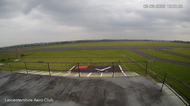 A view from a webcam at Leicestershire Aero Club showing an overcast sky with grey clouds, a grassy airfield with runways in the background, and a small helicopter landing pad with a white and red marking in the foreground. The time and date stamp in the upper right corner indicates it was taken on September 6, 2025, at 12:00:28.