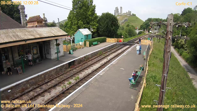 A small train station platform with a stone building on the left, where a couple of people are standing. There are two sets of railway tracks running through the scene, with some greenery and trees on both sides. A group of three people is standing near the far end of the platform, and a woman is sitting on a green bench closer to the camera. In the background, there is a hill with the ruins of a castle at the top, surrounded by lush green trees and grass. An old wooden utility pole with wires extends along the right side of the image, with green grass and a pathway beside it. The sky is partly cloudy.