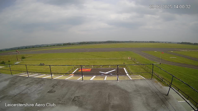 A view from a webcam at Leicestershire Aero Club showing a cloudy sky over a grassy airfield with runways in the background. In the foreground, there is an aircraft parking area with a small painted airplane symbol on the ground. A fence surrounds the parking area, and a windsock is visible on the left side. The timestamp indicates it is 14:00 on September 6, 2025.