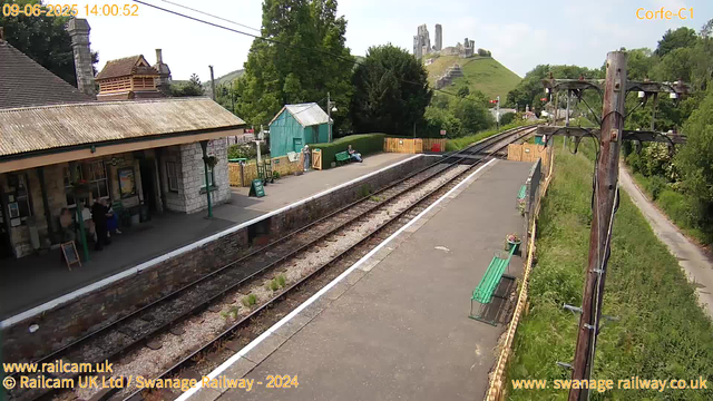 A small train station platform with a building on the left, featuring a stone exterior and a tiled roof. A few people are standing near the ticket machines. Two green benches and a person sitting on one are visible on the far side of the platform. There is a green shed and some colorful fencing near the benches. The railway tracks run through the center of the image, with a telegraph pole on the right side supporting wires. In the background, a grassy hill with ruins of a castle or similar structure is visible, along with lush green trees. The sky is slightly cloudy with some sunlight. The timestamp indicates the date as September 6, 2025, at 14:00.
