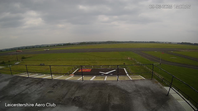 An overcast sky above a small airport runway with a windsock and a windsock with a flag nearby. Two small airplanes are visible in the distance on the runway. The foreground shows a rooftop and a helipad with a large white "X" and a red square, surrounded by a railing. The bottom left corner displays the text "Leicestershire Aero Club".