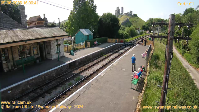 A small train station platform on a sunny day with a few elderly people sitting on benches, a man in a blue shirt standing, and a couple of people further along the platform near the fence. There are railway tracks running alongside the platform, a stone station building with a sloped roof on the left, and a green shed further behind. In the background, a hill with the ruins of an old castle or fortress atop it. A wooden utility pole with wires is on the right side of the image. Trees and greenery surround the area.